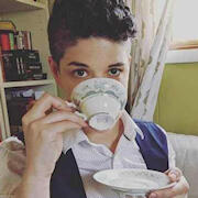 Individual with short, dark hair, dark eyes, pale skin and freckles. They are sipping from a teacup, which obscures much of their face. They are dressed in a white button-up shirt and a navy blue vest and are seated in front of full bookshelves.
